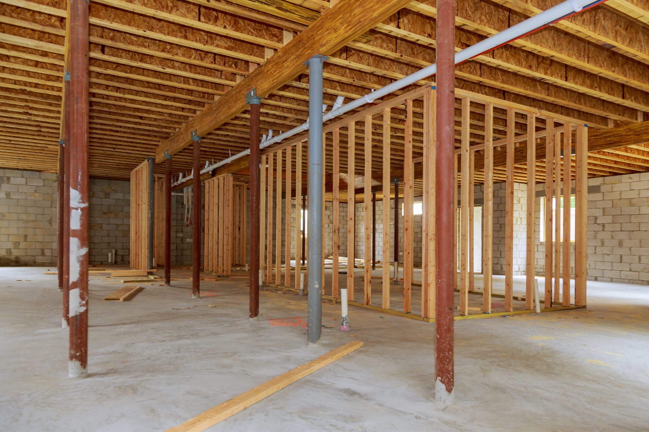An unfinished basement under construction with exposed framing, concrete walls, and plumbing pipes.